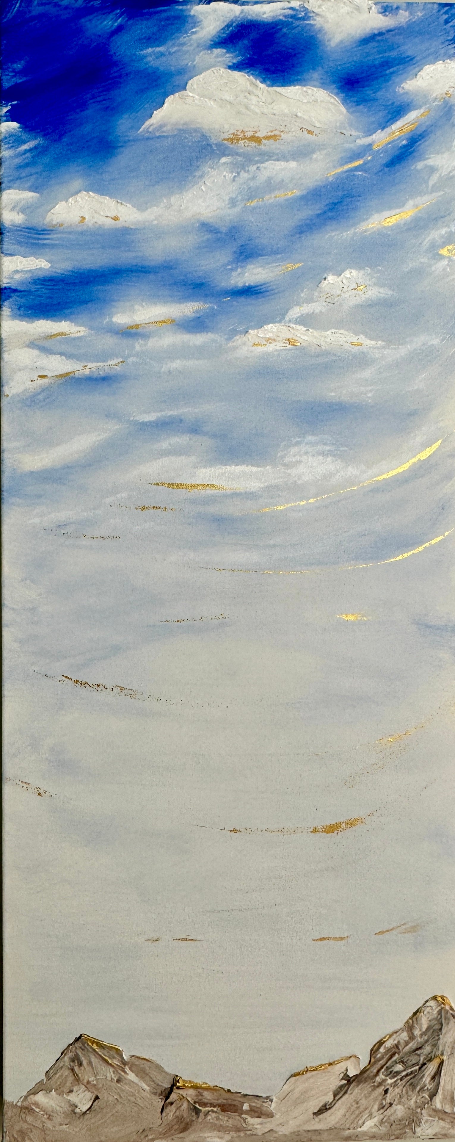 A tall vertical painting with a blue sky and clouds with whisps of gold, over a beige rocky mountain range.
