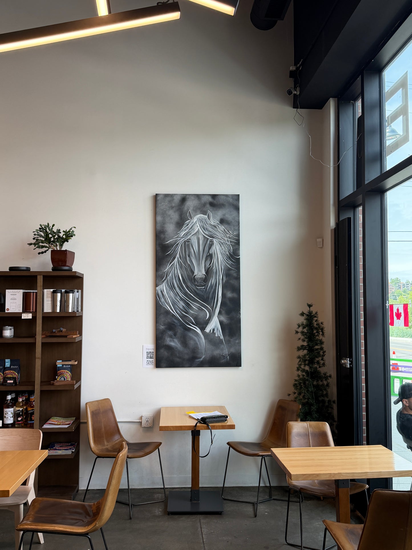 A vertical black and white canvas painting of a graceful horse portrait, displayed on a white wall in a modern business interior setting with wooden tables, chairs, and a bookshelf.