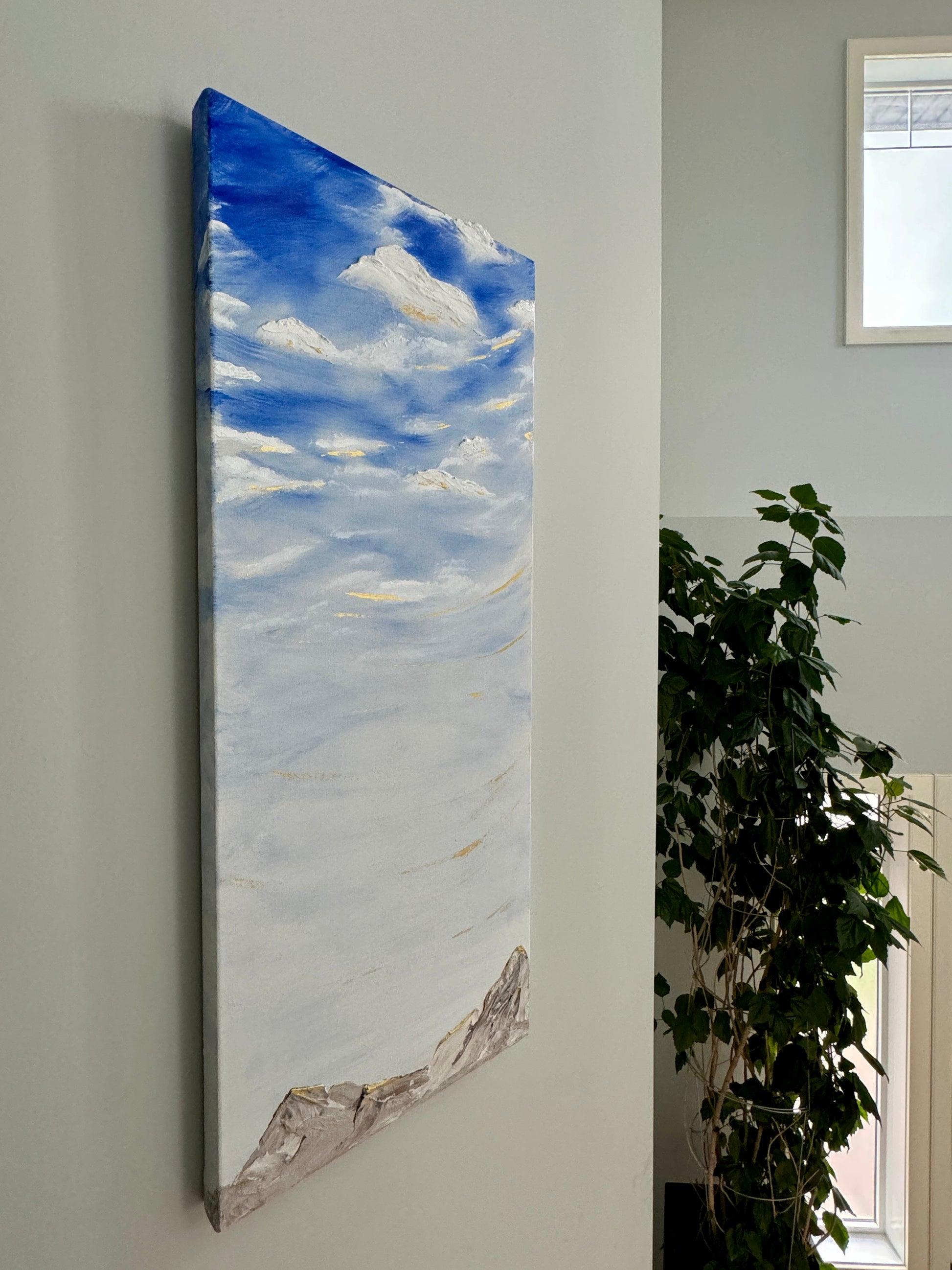 A tall vertical painting with a blue sky and clouds with whisps of gold, over a beige rocky mountain range.